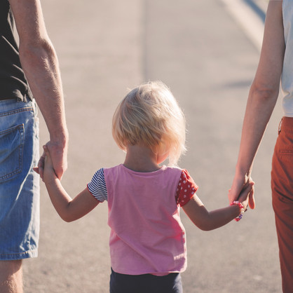 Rückansicht eines blonden Kindes, welches zwischen seinen Eltern läuft, ein Elternteil jeweils an einer Hand.