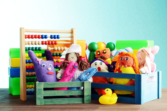 Sprachtherapeutisches Ambulatorium Eine Nahaufnahme verschiedener Spielzeuge in kleinen bunten Holzkisten. Man sieht Stoffpuppen, eine Gummiente, Spielfiguren und einen Abakus mit bunten Holzperlen.