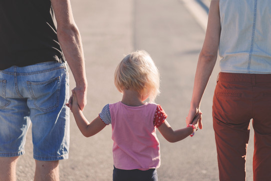 Psychologische-Pädagogische Ambulanz Rückansicht eines blonden Kindes, welches zwischen seinen Eltern läuft, ein Elternteil jeweils an einer Hand.