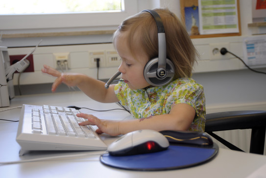 UK-Netzwerk Aufnahme eines blonden Kindes mit einem Logitech-Headset auf dem Kopf, welches an einer weißen Tastatur sitzt. Das Kind ist mitten in der Bewegung des Tippens und wirkt sehr konzentriert.